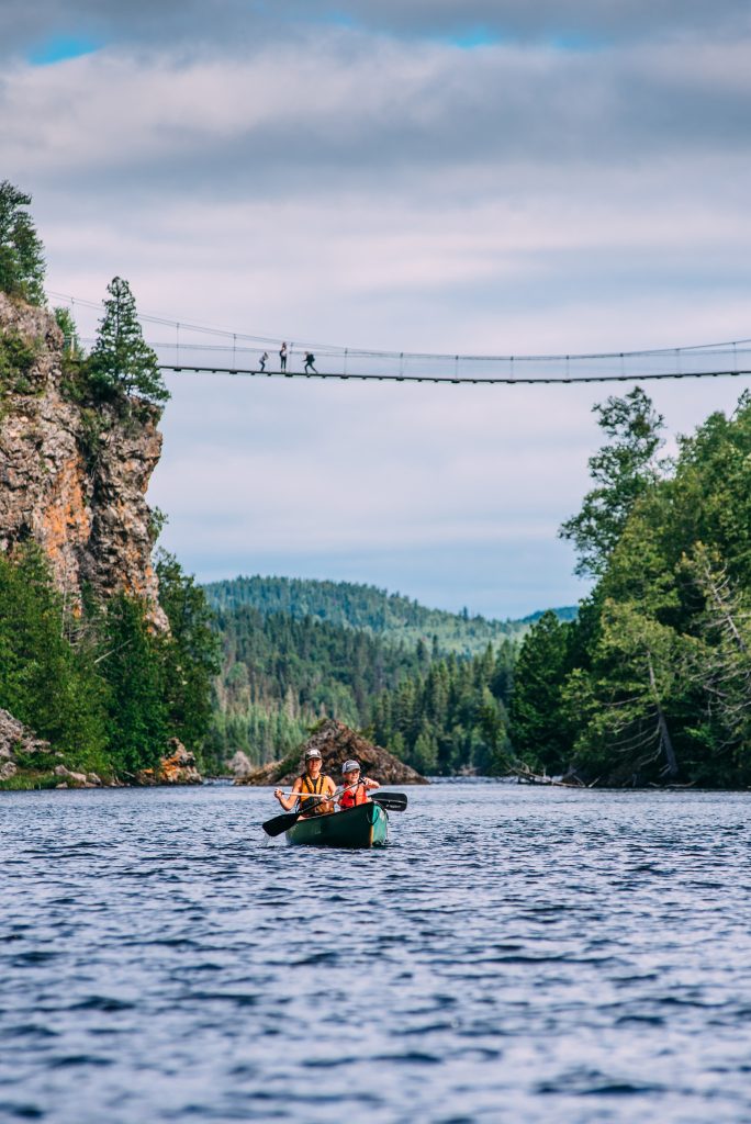 Deux personne en canot sur un lac au par national d'Aiguebelle. On loin, sur le pont suspendu, trois personnes.