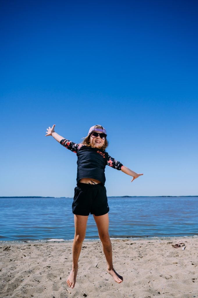 Sur une plage de sable, une jeune fille saute dans les airs avec ses bras et ses jambes bien tendus. Elle porte un costume de bain, des lunettes de soleil et une casquette avec la palette par en arrière. La jeune fille est tout sourire. Aller à la plage est une belle activité à faire en famille en Abitibi-Témiscamingue. 