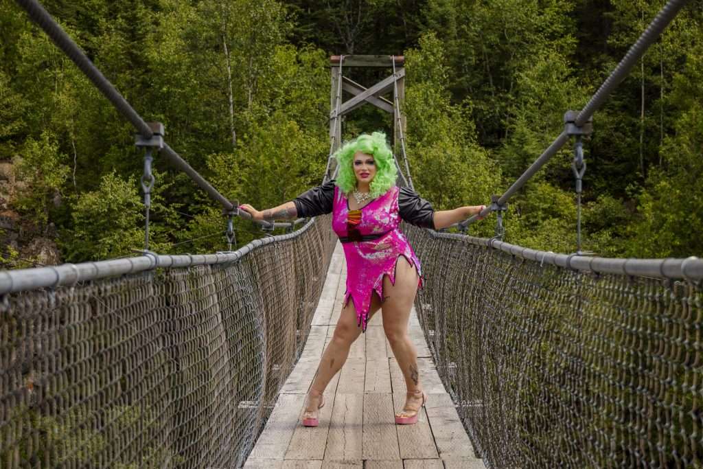 Sasha Baga, une dragqueen portant une robe très courte rose à paillette et une chevelure bouclée et vert fluo se trouve au milieu d'un pont suspendu. Elle nous fait face, ses main sont posées sur les câbles et elle prend un pose. Sasha porte des sandales à talons transparents et très hauts.