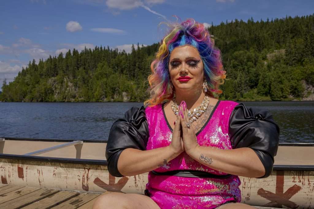 Sasha Baga, une dragqueen à la chevelure bouclée et multicolore est assise en tailleur et ses mains sont jointes, comme pour faire une prière. Elle a les yeux fermés et semble très zen.