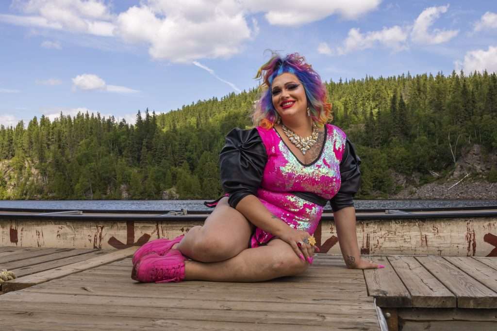 Sasha Baga, une dragqueen portant une robe très courte rose à paillette et une chevelure bouclée multicolore se trouve sur un quai, près d'un rabaska. Elle est assise et semble décontractée. Elle nous sourit. 
