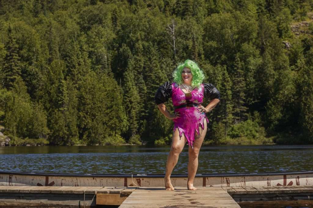 Sasha Baga, une dragqueen portant une robe très courte rose à paillette et une chevelure bouclée vert fluo se trouve debout sur un quai, près d'un rabaska.  Ses mains sont portées sur ses anches et elle nous sourit.