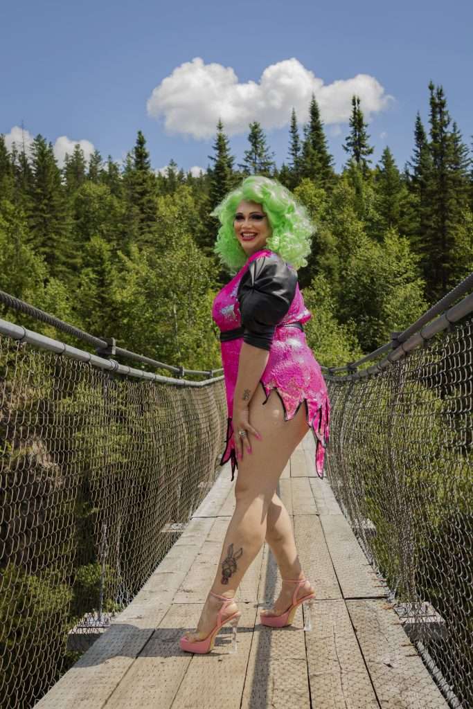 Sasha Baga, une dragqueen portant une robe très courte rose à paillette et une chevelure bouclée et vert fluo se trouve au milieu d'un pont suspendu. Elle est à de profil, mais nous regarde droit dans les yeux et elle nous sourit. Sasha porte des sandales à talons transparents et très hauts.