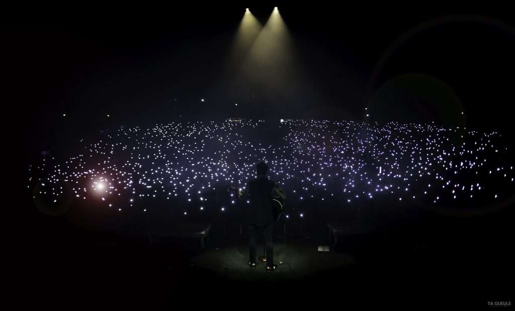 La photo présente une scène sur laquelle un chanteur est en prestation en soirée En arrière plan, on voit la lumière des téléphones des festivaliers.