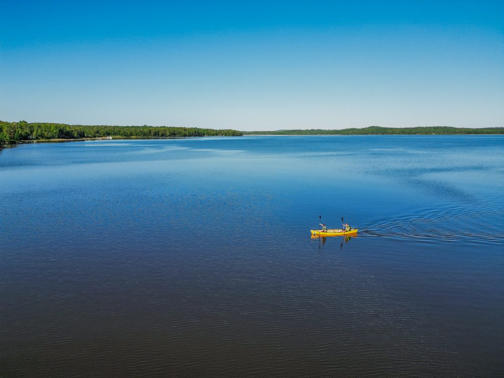 Sur un grand lac du témiscamingue, le lac des Quinze, on peut voir un canot jaune ainsi que deux personnes qui rament. Le ciel et bleu , la ligne d'horizon est une mince ligne verte d'une forêt lointaine et le lac est tout aussi bleu que le ciel.
